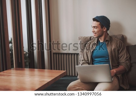 Portrait of good-looking guy in shirt sitting with notebook on his laps and looking away with smile