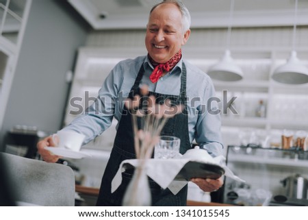 Low angle portrait of smiling gray-haired gentleman holding tray and serving cup of coffee. He wearing elegant red neckerchief