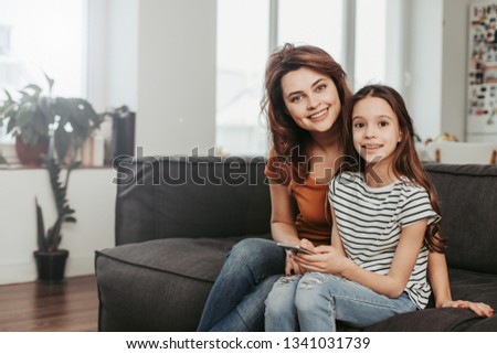 Close family relationship. Waist up portrait of happy smiling woman and her female child looking at camera while sitting on sofa at home with mobile phone. Copy space on left