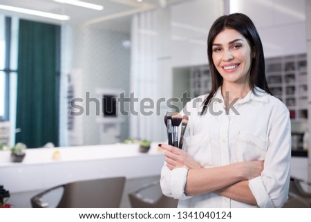 Half length of smiling lady standing in beauty saloon while holding brushes for visage. Beauty concept