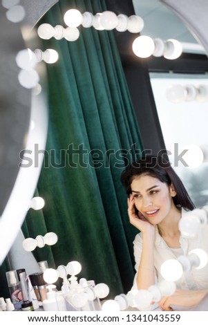 High angle of young charming lady looking at mirror while leaning on her elbow. Bottles with cosmetics situating on table near female hand. Beauty concept