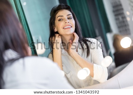 Reflection in mirror. Smiling pretty lady in bathrobe posing for camera and sitting at mirror in hairdressing saloon. She is touching her neck while admiring herself. Focus on happy face