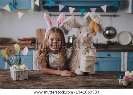 Preparing for Easter. Waist up portrait of happy smiling female child staying on festive decorated kitchen wearing rabbit ears and holding rabbit toy with colored eggs