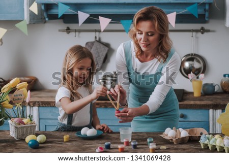 Preparing for Easter. Waist up portrait of happy woman and her female child coloring eggs on festive kitchen at home