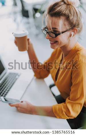 Cheerful young woman in an orange blouse sitting at the office table with wireless earphones in her ears and smiling while enjoying her coffee
