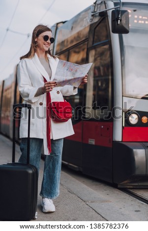 Full length portrait of charming lady in white trench coat searching route on map. She standing near trolleybus and smiling