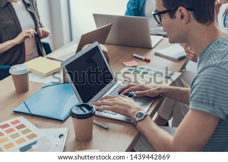 Cropped portrait of man in glasses sitting at the office desk with laptop while colleagues sitting on background. Focus on laptop screen