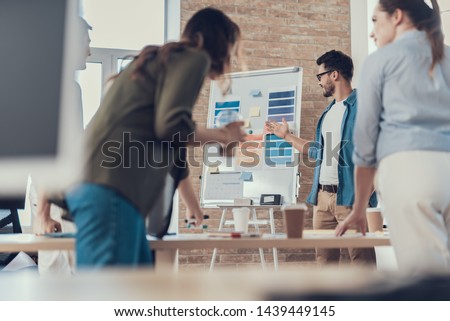 Waist up portrait of coworkers standing at the office desk and debating. Enjoyed man showing documentation on whiteboard