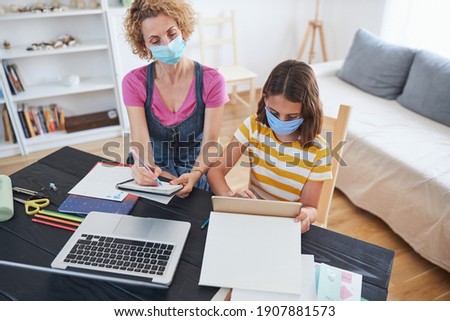 Child home studying education, homeschooling, with private tutor mother with protective mask in the time of viruses, flu and seasonal pandemic.