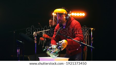 Male sound designer in protective face shield using hammer to break glass jar near microphones and recording soundtrack for movie near colleague in studio