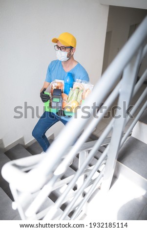 Delivery guy on the building staircase with protective mask holding box with groceries and POS for contactless payment.