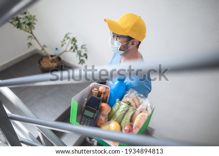Delivery guy on the building staircase with protective mask holding box with groceries and POS for contactless payment.