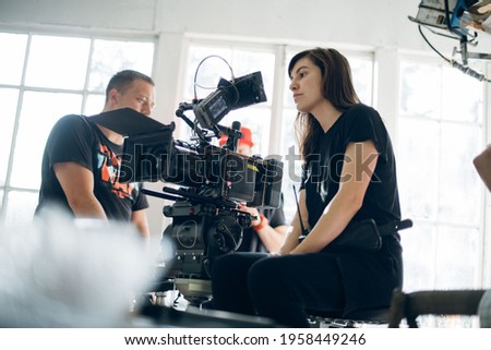 Female director of photography with a camera on a movie set. Professional videographer on the set of a movie, commercial or TV series. Filming indoors, studio