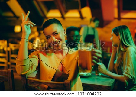Young happy African American woman having fun with friends during the night out in a pub and toasting with beer while looking at camera.