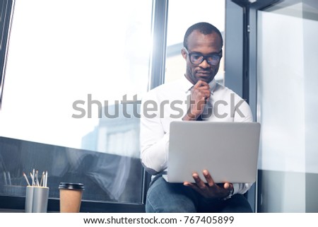 Searching for solution. serious man is sitting on table in office. He is looking at screen of modern laptop with concentration while touching his chin. Copy space in the left side