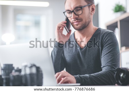 Portrait of happy unshaven man chatting by mobile while working with notebook computer in apartment. Conversation concept