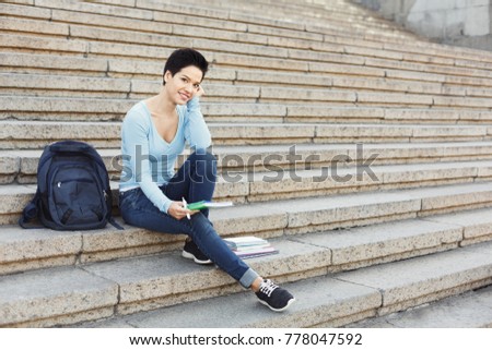 Young smiling female student making notes in notebook, sitting on university stairs. Education, inspiration and remote working concept.