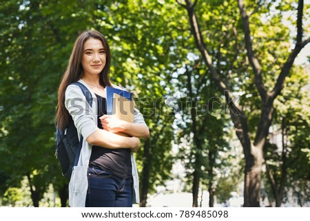 Attractive kind student girl with books in park outdoors, smiling at camera, dreaming or thinking during break, having rest in campus. Education concept, copy space