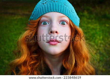 Surprised red-haired girl in a blue hat with eyes wide open looking directly into the camera