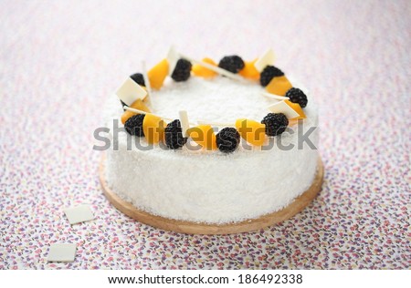 White Marshmallow Cake with coconut flakes, blackberries and pieces of peach, on a wooden cutting board and on a light tablecloth.