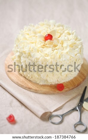 White Forest Cake - White Chocolate Cherry Cake on a wooden cutting board and scissors, on a light beige background.
