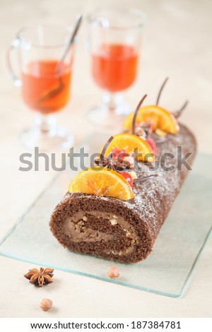 Chocolate Swiss Roll Cake with candied orange slices, cherries and hazelnuts, two cups of fruit tea on a light beige background.