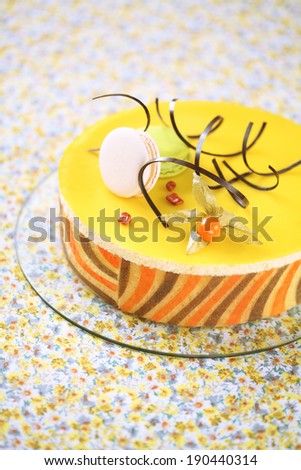 Tropical Mousse Cake (Charlotte) decorated with macarons, physalis and chocolate spirals, on a yellow floral tablecloth.