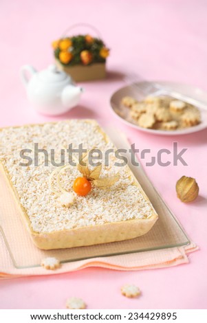 Pear and Coconut Tart decorated with Physalis on a glass cutting board, little teapot and little cookies, on a light pink background.