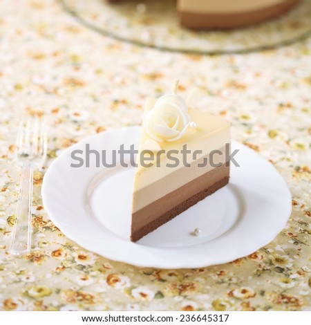 Piece of Three Chocolate Mousse Cake on a white plate, on a light yellow floral table cloth.