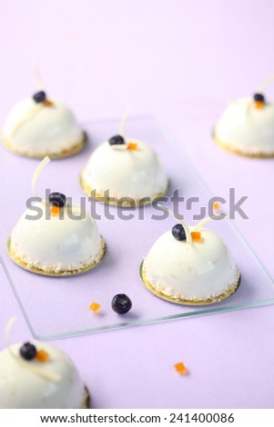 Blueberry and Orange Mousse Cakes decorated with white chocolate curls and toasted coconut flakes, on a light purple background.