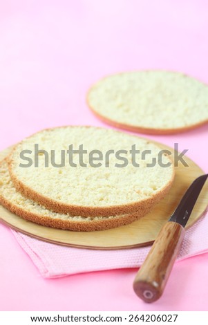 Three layers of Biscuit Sponge Cake on a wooden cutting board and a knife, on a light pink background.