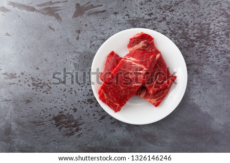 Top view of several pieces of beef chuck boneless short rib grilling steaks on a white plate on a gray counter top illuminated with natural lighting.