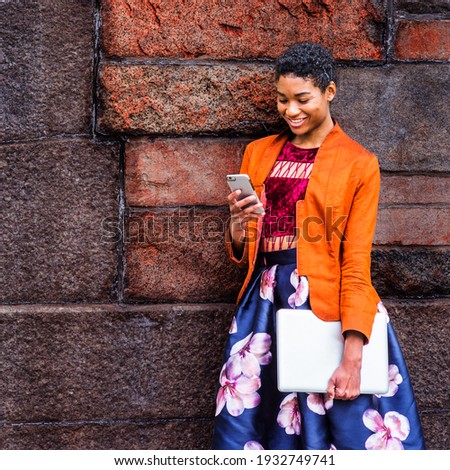 A Young African American Woman is texting on a cell phone outside, wearing an orange, red jacket, a flower-patterned skirt, carrying a laptop computer, standing by a stone wall on campus, smiling.