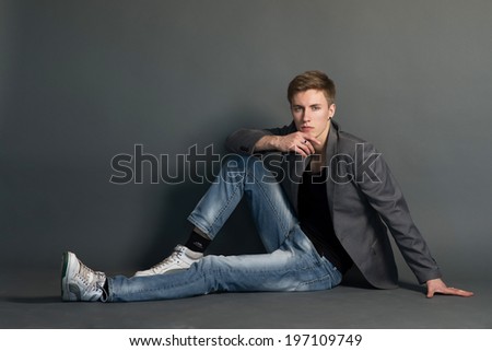 Man in jeans and jacket, sitting. Dark grey background