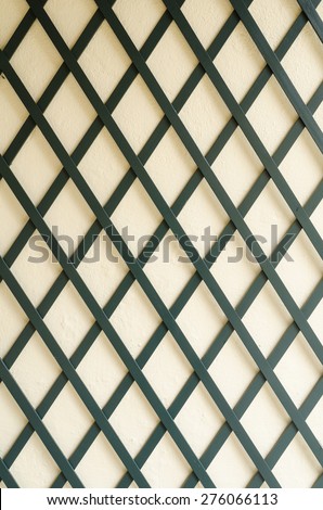 dark thin wooden boards forming a regular diamond pattern in front of a bright cement wall.