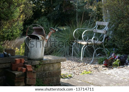 Rural Country Garden scene containing rusty watering can, garden bench, garden wall, gravel path, patio etc.