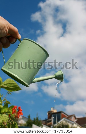 A small hand held watering can being held aloft against an urban garden scene.Water emerging from spout of watering can.