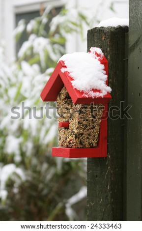 A wooden bird feeder, nailed to a green wooden post, filled with nuts and seeds,covered with fresh crisp white snow