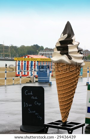 A plastic Ice Cream Cone next to a hand written black chalk board with the text \'Cod & Chips £5 served all day\'. Located on Weymouth pier in Dorset UK outside a cafe.