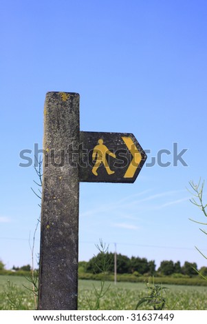 Public footpathsign set on a concrete post with countryside backdrop on a clear blue summer sky.