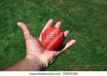 A red leather cricket ball caught in an open hand. Set over a green grass area.