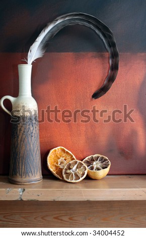 Still life image of dried fruit. A lemon and Orange set on a wooden mantle shelf with a ceramic pot and curling feather. All set against a grunge styled background.