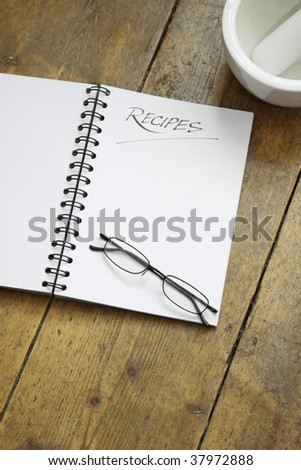 A blank wire spiral bound recipe book with the title 'recipe' hand written at the top of the page. A pair of black framed glasses rest on the page. Set on a wooden kitchen table top.