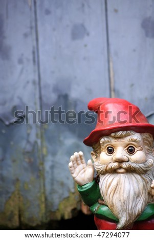 A small bearded garden gnome waving his hand. Set against a soft focus wooden garden shed door.
