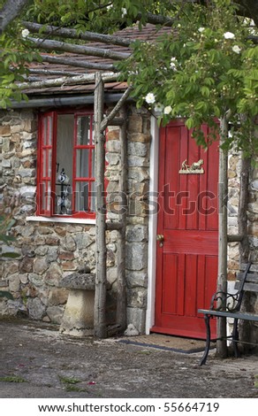 A stone constructed summer house with a terracotta slated roof, a red painted wooden door and window frames. A wooden trellace coverd in white roses adorns the front. Located in Devon,England.