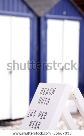 A hand painted sign advertising beach huts for hire. Blue and white beach huts visible in soft focus to background. Located at Beer, in Dorset, England.