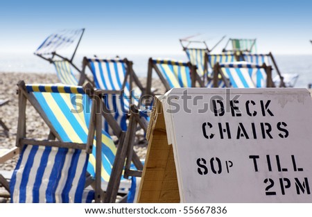 A landscape format image of a hand made advertising board, advertising beach side deck chairs for hire. Various colored deck chairs in soft focus to background. Located in Beer in Dorset, England.
