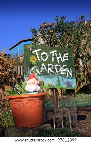 A portrait format image of a terracotta coloured flower pot and garden gnome in front of a garden fork and hand painted wooden sign with text spelling \'to the garden\'.