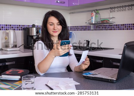 Happy young woman with laptop using credit card and phone in kitchen at home