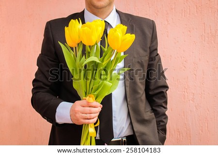 A man wearing a business suit, holding a bouquet of yellow tulips. The man gives a bouquet of flowers.